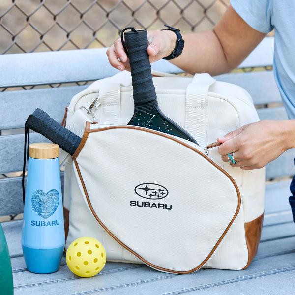 Photo of a cute blue Subaru heart bottle next to a pickleball set with a woman's hand taking a raquet out of the bag